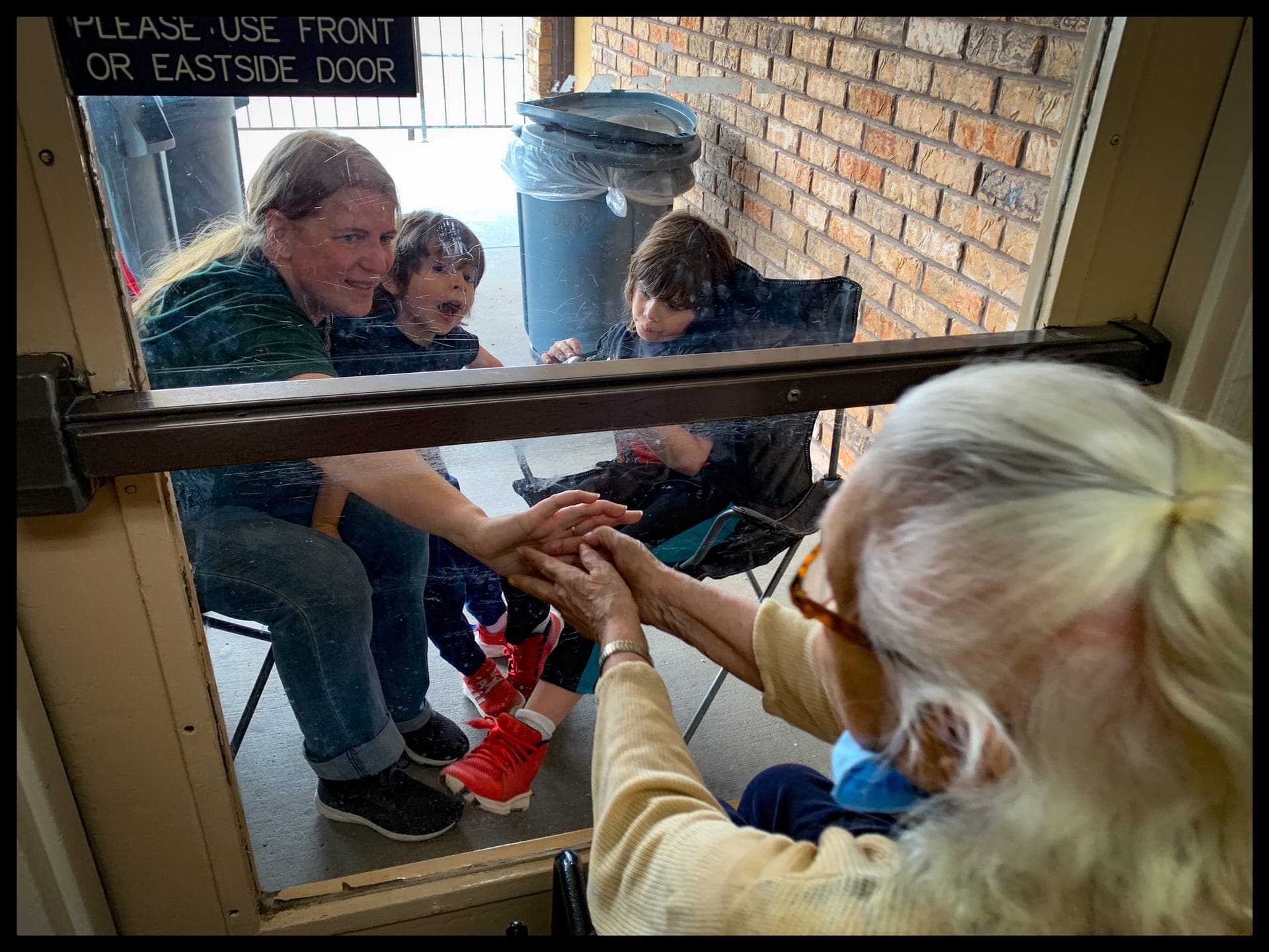 A family outside a window waves to an older woman inside a nursing home