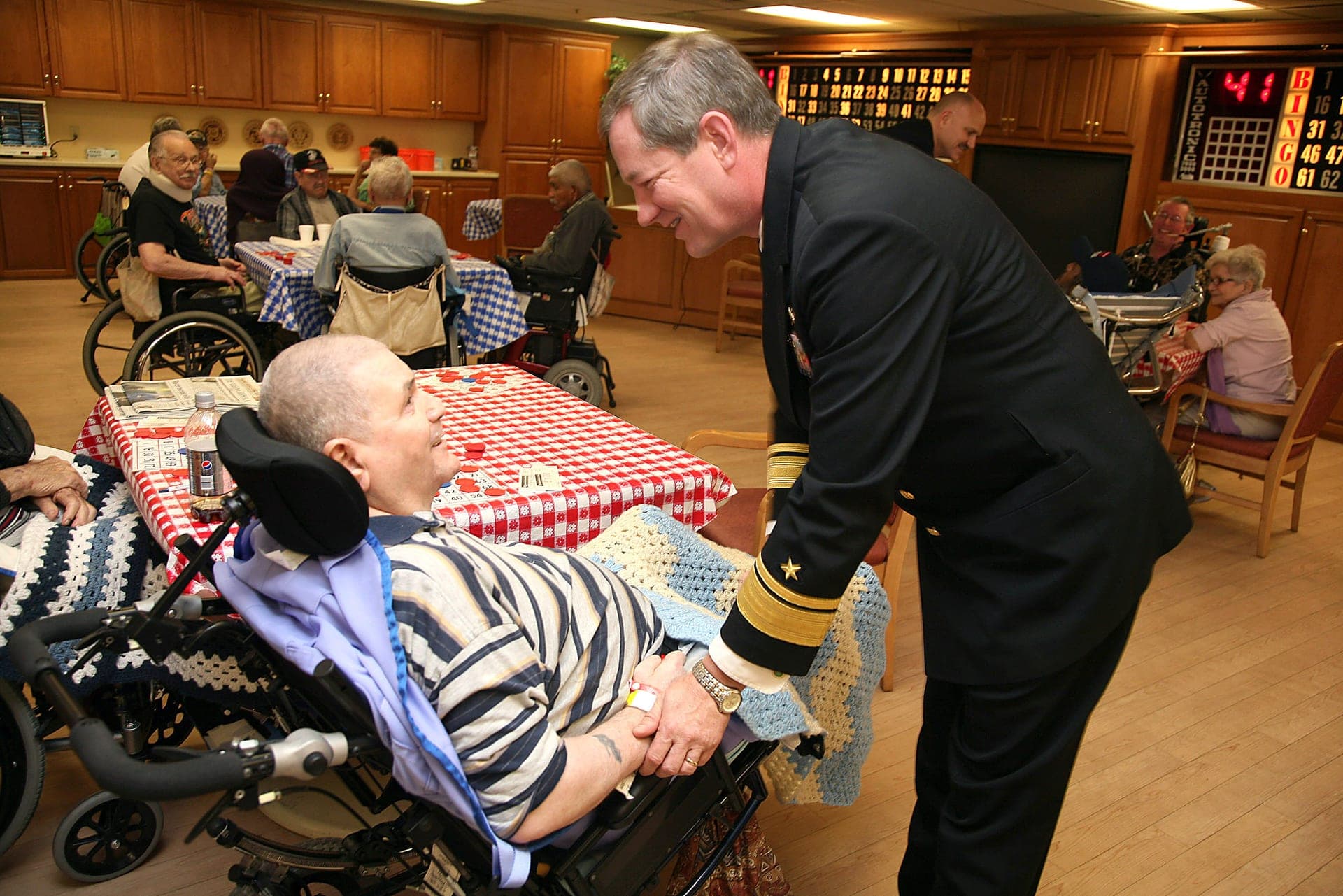 A U.S. Navy rear admiral shakes hands with an older woman while other residents sit nearby in a nursing home day room