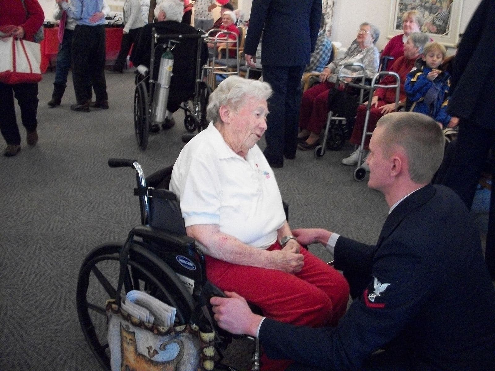 A Coast Guard crewman sits beside an older woman and talks with her in a Nantucket nursing home room